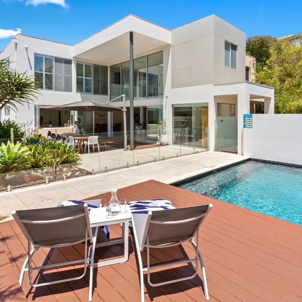 Two deck chairs on a patio next to a pool, looking onto a modern two-storey house.