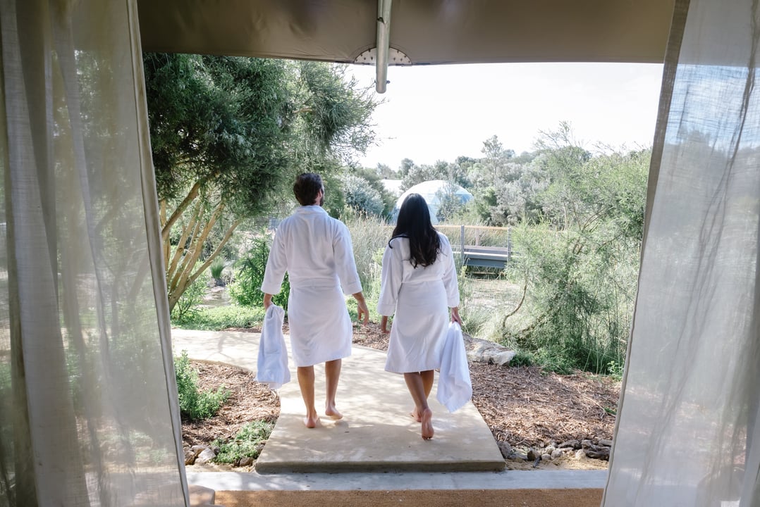 A couple hand in hand walking out of their Glamping tent in white robes