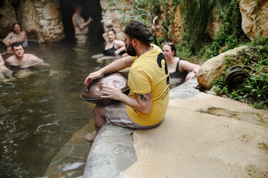 A person playing the hand pan with their legs dangling in hot springs water