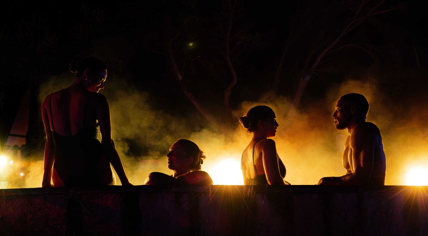 A group of four people sitting on the edge of a hot spring with chakra-inspired lighting glowing on them as part of the Seek the Heat winter campaign