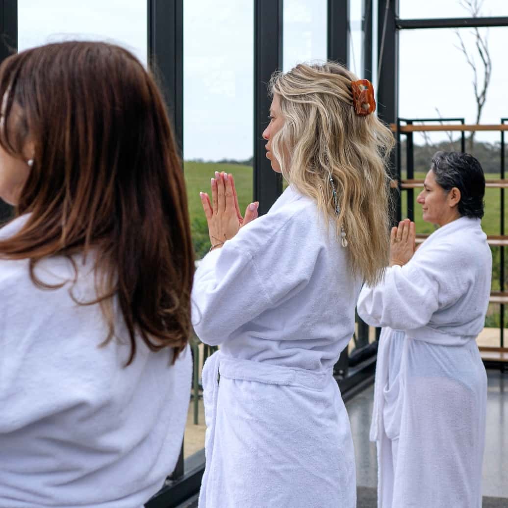 Three people standing in white robes with their hands in a prayer position during the Stretch and Breathe class