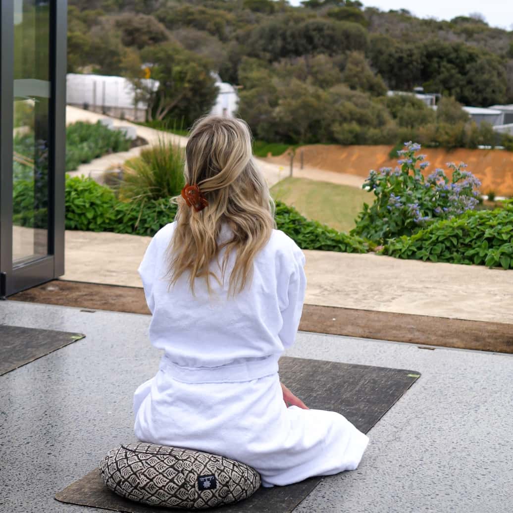 A woman in a white robe is sitting on a round cushion during a Stretch and Breathe class
